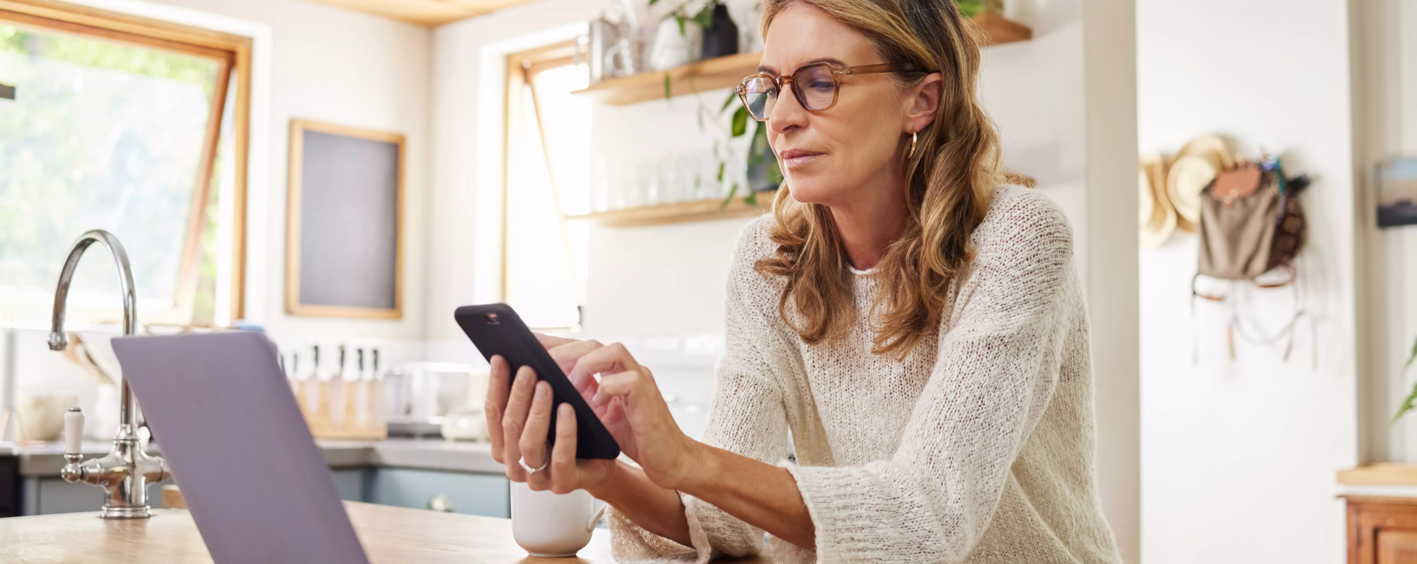 Femme, entrain de chercher sur son téléphone devant un ordinateur portable sur ilot de sa cuisine.